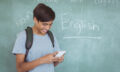 Happy schoolboy with backpack using mobile phone in classroom