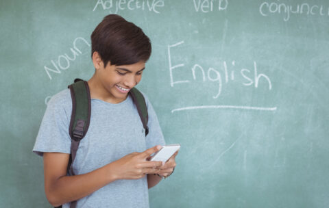 Happy schoolboy with backpack using mobile phone in classroom