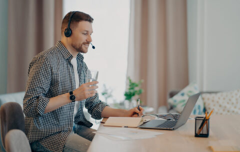 Male college student listens academic podcast sits at desk with laptop consults client by video makes notes drinks water from glass watches webinar dressed in checkered shirt poses at home office