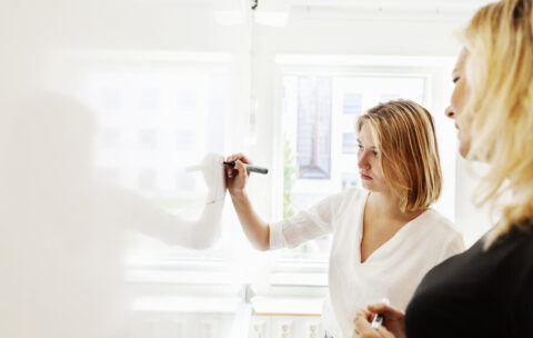 Young woman writing on whiteboard while standing by classmate in classroom